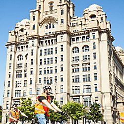 People wearing orange high vis jackets and helmets ride electric bikes in front of the Liver Building.