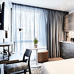 the view across a modern apartment. The room is light with white and grey details including a small kitchen area, large window and floor length curtains and a wall mounted television.