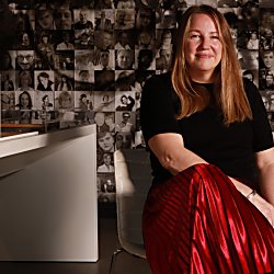 An image of a woman sitting on a chair with her leg crossed, wearing a red skirt and black top.