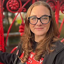 A woman looking towards the camera wearing black glasses and a black jacket with red gates behind her.