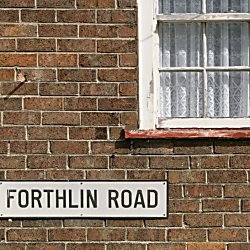 A brick wall on a house with a window to the top right. There is a drain pipe to the left with a sign for Forthlin Road.