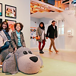 A girl and a boy sit smiling on a large, soft, dog-shaped seat in a brightly lit, family-friendly exhibition space. Behind them, photos of animals hang on the wall. In the background, two adults walk through the room, observing the displays.