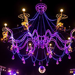 A large chandelier lit up in purple lights with performers on it as part of an act.