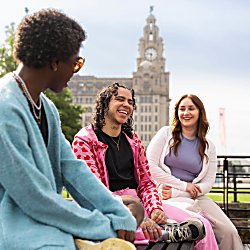 3 people sit on a bench on Liverpool Waterfront with the Royal Liver Building in the background