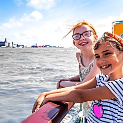 Two young girls stand on the Mersey Ferry smiling on a sunny day