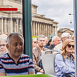 A group of people on the top deck of an open top bus tour of Liverpool. In the background the large neo-classical St George's Hall building can be seenn