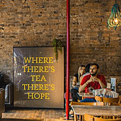 Inside the LEAF cafe. Against an exposed brick wall is a large print that reads 'where there's tea there's hope.' People are sitting at wooden tables and chairs in various chalky colours. There's also a disco ball hanging from the ceiling and a large red supporting column.