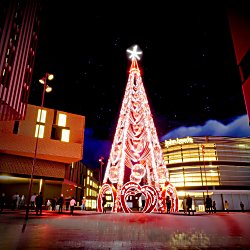 Illuminated Christmas tree installation in Liverpool ONE at night, featuring glowing red and white festive lights, decorative arches and a bright star at the top. Visitors walk through the plaza surrounding the display, with the John Lewis building and city centre architecture lit in the background beneath a dark, starry sky.