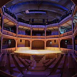 An image of the cockpit in court theatre style at Shakespeare North Playhouse. The image is dark with central spotlight on centre stage. There is wooden beams surrounding the cockpit and stage.