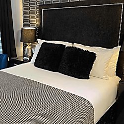 A plush double bed with white duvet covers and two square black pillows on top. The bed has a black headboard with silver studs outlining the square headboard and along the bed features a houndstooth throw.