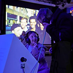 A child and an adult standing listening to something with headphones on, there's a large digital screen behind them.