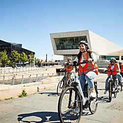 A group of people on electric bikes wearing orange high vis jackets on Liverpool's pier head.