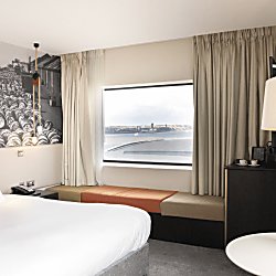 A light, spacious hotel room with a large bed with white sheets. Behind the bed is a black and white historic image of Liverpool Docklands. There's a cushioned bench style seat underneath a window, with beige curtains open.