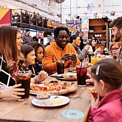 A family are sitting at bench tables in a food market. There are different coloured food vendor stalls in the food market.
