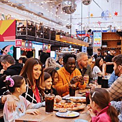 Families and friends at a street food market. There's glitter balls hanging from the ceiling and bright coloured food vendor windows.