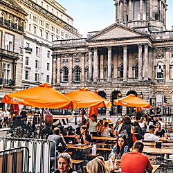 Liverpool Town Hall in the background of a shot of people eating a drinking at tables on a street. There are large orange umbrellas next to some tables.