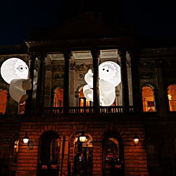 Two large glowing inflatable figures with cartoon-style eyes appear suspended between the columns of a grand historic building at night. The illuminated white sculptures contrast against the dark sky, while warm orange light shines through the building’s windows, creating a playful and dramatic light installation.