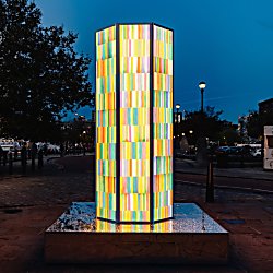 A tall illuminated tower made up of colourful vertical light panels glows on Liverpool’s waterfront at dusk. Standing on a reflective base, the geometric sculpture shines in shades of yellow, green, pink and blue, creating a vibrant focal point against the evening sky and surrounding riverside setting.