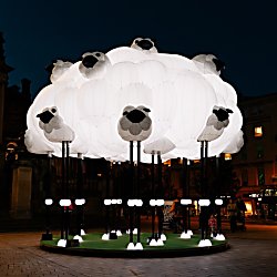 A large illuminated sculpture resembling a flock of sheep forms a glowing white cloud in a city square at night, with individual sheep heads and legs extending from the installation.