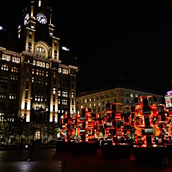 A striking red illuminated sculpture made of stacked geometric forms glows in a city square at night, set against the iconic Royal Liver Building and surrounding historic architecture. The artwork's reflective surfaces create a vivid contrast with the dark sky and warmly lit waterfront skyline.
