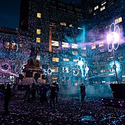 Visitors gather in a city square at night surrounded by illuminated flower-shaped light sculptures. Bright blue, purple and white projections fill the space with sparkling patterns and beams of light, creating an immersive outdoor artwork against a backdrop of historic buildings and glowing windows.