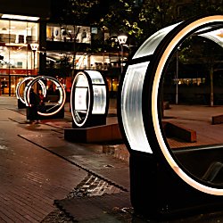 A series of large illuminated circular installations line a modern city plaza at night, glowing with animated digital displays and creating an interactive light trail against a backdrop of glass-fronted office buildings.