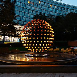 A glowing circular light sculpture made up of dozens of illuminated bulbs radiates from a central hub above a reflective water feature. Set within a landscaped urban plaza at dusk, the artwork stands in front of a curved glass-fronted building, with trees and soft lighting surrounding the installation.