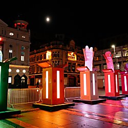 A colourful light installation features a row of illuminated gift boxes in a city square at night, each with a glowing object emerging from the top. Historic buildings and shopfronts are lit in the background, while vibrant green, orange, pink and red lights reflect across the pavement.