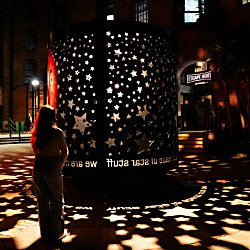 A large cylindrical light installation decorated with star-shaped cut-outs glows in an urban courtyard at night. Light shines through the stars, casting intricate patterns across the surrounding pavement, while a visitor stands nearby observing the artwork against a backdrop of brick buildings and illuminated walkways.