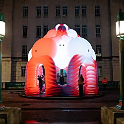 A large illuminated inflatable sculpture glows in shades of red, pink and white in a city square at night. Visitors stand beneath its arched structure, while historic buildings and tall street lamps frame the artwork against the dark sky.