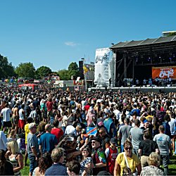 Wide outdoor festival scene with a large crowd standing on grass in front of a main stage. The stage features lighting rigs, banners, and a large central screen, with trees and fairground rides visible in the background under a clear blue sky.