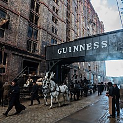 A large brick building with a sign going between another building that says Guinness on it. 

There is a horse and cart walking through the middle of the street.