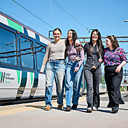 Two people walk along a railway platform beside a London Northwestern Railway train. The train features a blue, white and green livery, while overhead power lines and station infrastructure are visible in the background. The two passengers walk arm-in-arm along the platform, with clear blue skies overhead.