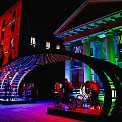 Purple and green lights illuminating the area with people on bikes as part of the installation.