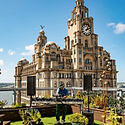 A person standing on a rooftop with the Royal Liver building behind them, they're playing music and the sky is blue.