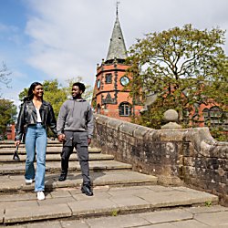 Two people walking down stairs over a bridge, they are walking with a clock tower behind them in the distance. There is trees to the right of the image.