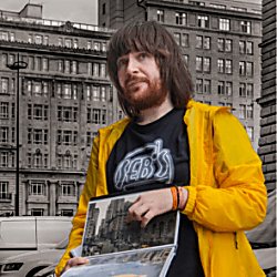 A man standing with a booklet showing a film location. He is wearing a bright yellow coat and pointing to the photo.