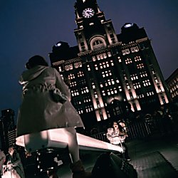 A seesaw lit up with The Royal Liver Building behind it, there's two people sitting on the seesaw swinging from side to side.