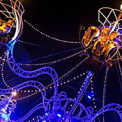 A large chandelier art piece with people sitting on it as part of a performance.