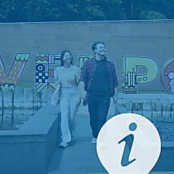 Two people walking towards the camera with a large Liverpool sign behind them. They are smiling brightly and the image has a blue wash over it with an I to the bottom right of the image.