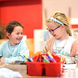 Two young children smiling and looking at each other, with a pot of pens in front of them.
