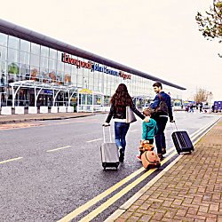 A man and woman are pulling small suitcase. There is a young child between them pulling a teddy bear suitcase. They are outside a modern, glass fronted building which is Liverpool Airport