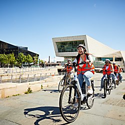 People riding bikes on Liverpool Pier Head wearing orange high vis jackets.