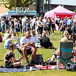 A group of people sitting on picnic blankets and chairs in a park at a food and drink festival.