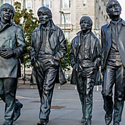 Bronze statue of four walking figures in a city plaza, each wearing coats and mid‑20th‑century clothing. One figure holds a coat while the others have hands in pockets. The sculptures stride forward side by side on a paved walkway, with a large stone building, trees, and modern city structures in the background under an overcast sky.