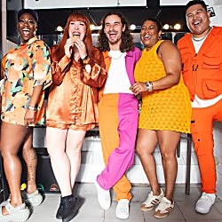 A group of five people stands in a brightly lit space with a black-and-white wall. They are all laughing and displaying energetic expressions. Each person is wearing colorful, bold outfits in orange, pink, and yellow tones. They appear to be enjoying a joyful moment together.