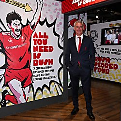 A Ian Rush stands inside a Liverpool Football Club museum exhibition beside a large graphic display celebrating the club's all-time leading goalscorer. The exhibition features bold red-and-white comic-style artwork, football memorabilia, trophy displays and interactive screens, with prominent text reading &ldquo;All You Need Is Rush.&rdquo;