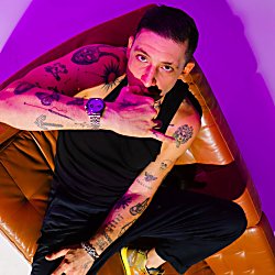 A studio portrait of a person sitting diagonally on a tan leather armchair, viewed from a slightly elevated angle. The person is wearing a black sleeveless top, black trousers, and yellow trainers. One arm rests across the lap while the other is raised, with the hand positioned near the mouth in a gesture resembling a phone call sign. The person has multiple visible tattoos on both arms, including various small designs and symbols. A wristwatch with a metallic strap is worn on one wrist, along with a bracelet.
The background is minimal and clean, with strong coloured lighting creating a gradient effect—magenta/pink tones dominate one side while a softer purple and white blend appears on the other. The lighting casts bold shadows behind the chair, adding contrast and depth to the composition.
The leather chair has a soft, slightly wrinkled texture, and its warm brown colour contrasts with the cool-toned lighting. No text is visible in the image.