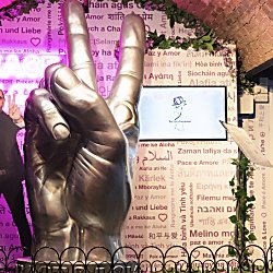 Large metallic sculpture of a hand making a peace sign displayed indoors beneath a brick arch, surrounded by decorative greenery and a patterned wall featuring the word “peace” written in multiple languages. A life-size cutout figure stands to the side, also holding up a peace sign. Two screens and informational plaques are positioned around the display, with pink and purple lighting illuminating the scene.