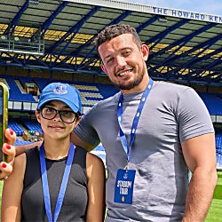 A man and a young girl are standing smiling and looking at the camera. The football stadium is behind them, with the young girl wearing a blue hat, and both have blue lanyards around their neck.